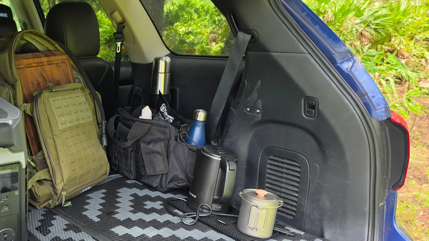 Road-trip coffee setup in an open SUV with travel gear, kettle, metal cup, and insulated bottle.